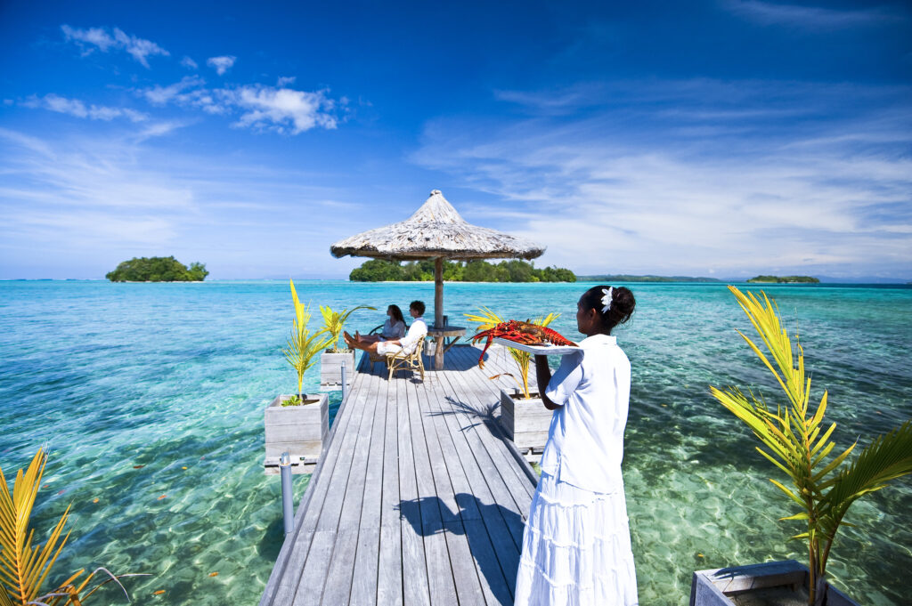 A couple enjoying a meal under an umbrella by the water in the Solomon Islands being served by a waitress